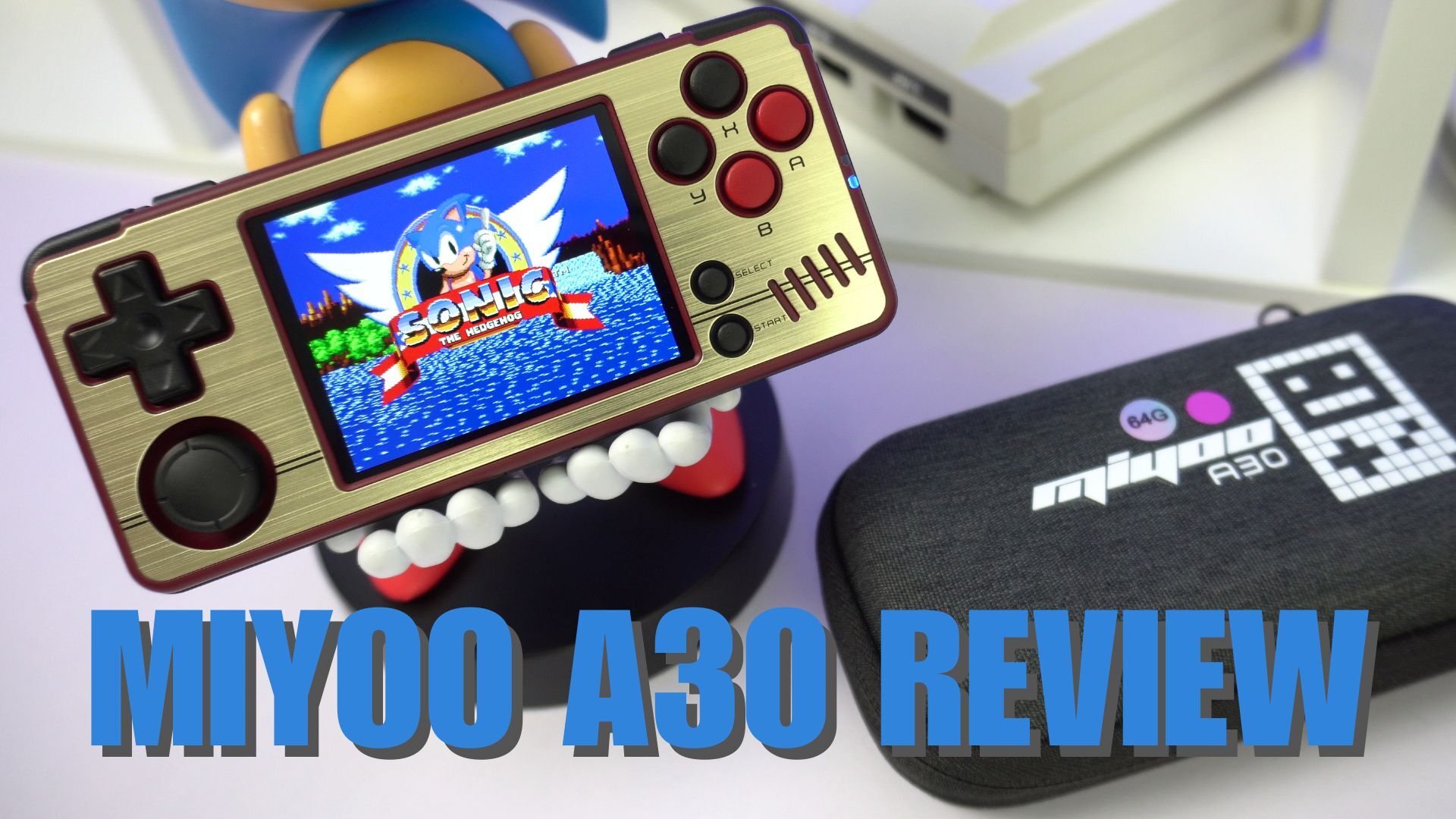 retro-style handheld gaming console, the Miyoo A30, is displayed prominently. Its gold and black design features a screen showing Sonic the Hedgehog, surrounded by classic gaming controls including a D-pad, circular buttons, and an analog stick. The device is held up by a playful figurine with large teeth visible. In the foreground, a black protective case bears the Miyoo A30 logo. The image showcases the compact size and nostalgic appeal of the console, emphasizing its portability and classic gaming capabilities. Bold blue text at the bottom reads "MIYOO A30 REVIEW," indicating the purpose of the image. The overall composition highlights the blend of modern technology with retro gaming aesthetics, appealing to enthusiasts of classic video games