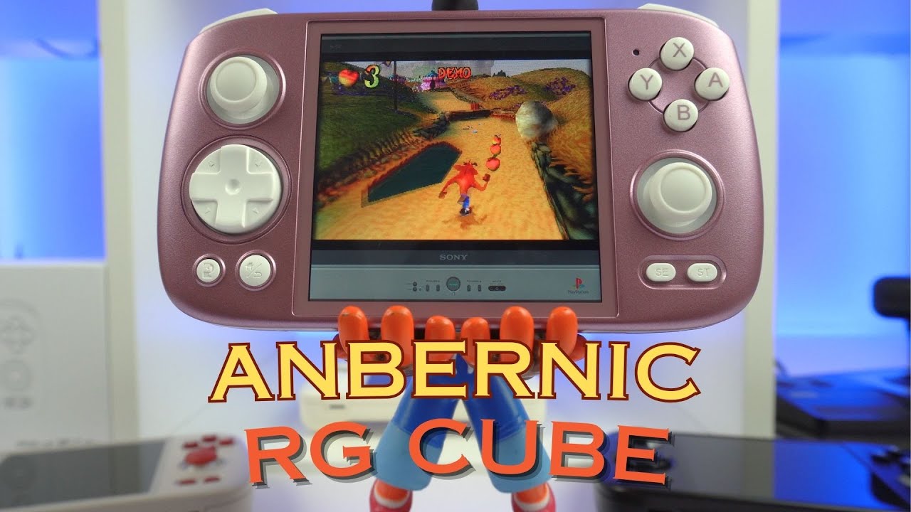 The image shows a handheld gaming console labeled "ANBERNIC RG CUBE" in the foreground. The console has a pinkish color and features a D-pad, two analog sticks, and several buttons, including X, Y, A, and B buttons. The screen of the console displays a gameplay scene from "Crash Bandicoot," where the character is running along a path collecting items. The console appears to be held up by a figure, possibly a toy or model, visible below the console. The background features a clean, modern setup with a blue-lit environment, and some other tech gadgets are partially visible.