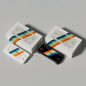Top-down view of white and black AYANEO Pocket ACE units placed beside their respective retail packaging, each with colorful branding and console imagery printed on the box lids.