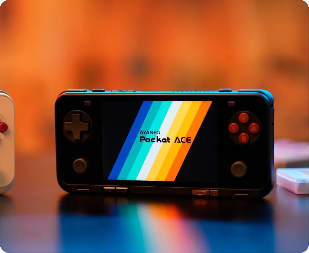 A handheld gaming console, the AYANEO Pocket ACE, displayed on a table with a colorful gradient logo on its screen. The device features a D-pad, analog sticks, and action buttons, all illuminated in a warm ambient setting with a blurred orange background.