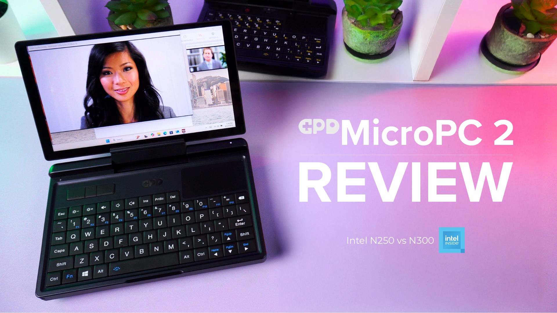 A dynamic thumbnail for a GPD MicroPC 2 review video. A black GPD MicroPC 2 is prominently featured in the foreground, open and angled towards the viewer, with its screen displaying a video conference call featuring a woman's face. The laptop's compact keyboard and trackpad are visible. The background is a white shelf with potted plants and another GPD MicroPC 2. The desk below the laptop is a vibrant pink and purple gradient. Large white text to the right of the laptop reads "GPD MicroPC 2 REVIEW", and smaller text below that says "Intel N250 vs N300" alongside the "Intel inside" logo.