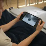 ONEXPLAYER 3 handheld gaming PC in hands on a soft bed, angled black console with glowing sticks and snowy driving game