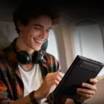 Smiling traveler on airplane holds black ONEXPLAYER 3 handheld gaming PC, angled rear view with textured vents